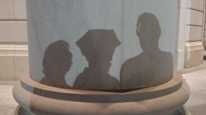 Shadow of Tanya Mikulas wearing 6-pointed PhD graduation tam with family on Shelby Hall pillar.
